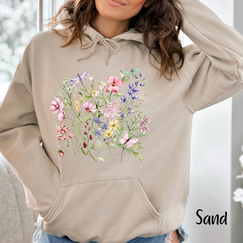 K&ouml;nnte beinhalten: Ein beiger Hoodie mit einem Blumenmuster mit rosa, gelben, blauen und gr&uuml;nen Blumen, Libellen und Schmetterlingen. Der Hoodie hat eine Kapuze mit Kordelzug und eine K&auml;ngurutasche. Das Wort "Sand" ist in Schwarz in der unteren rechten Ecke des Bildes gedruckt.