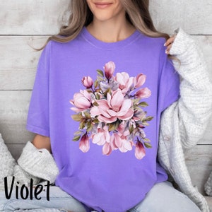 Peut inclure: Un t-shirt violet avec un motif floral compos&eacute; de fleurs de magnolia roses et de feuilles vertes.