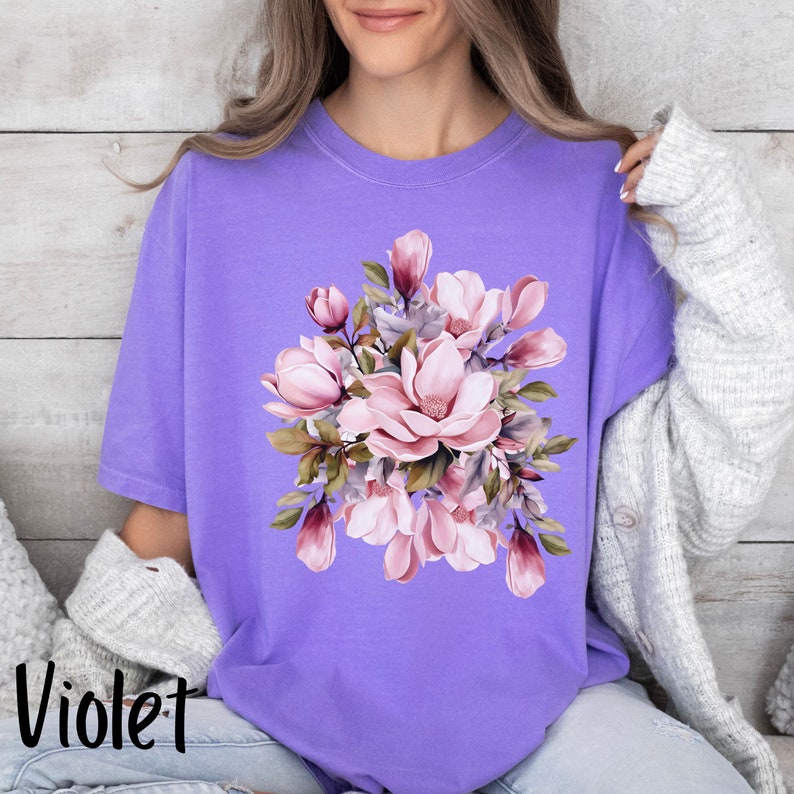 Peut inclure: Un t-shirt violet avec un motif floral compos&eacute; de fleurs de magnolia roses et de feuilles vertes.