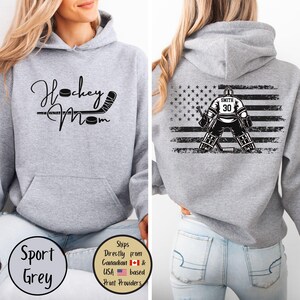 May include: Sport grey hoodie with "Hockey Mom" text and hockey stick graphic on the front. The back features a hockey goalie graphic with the American flag and the name "Smith" and number "30".