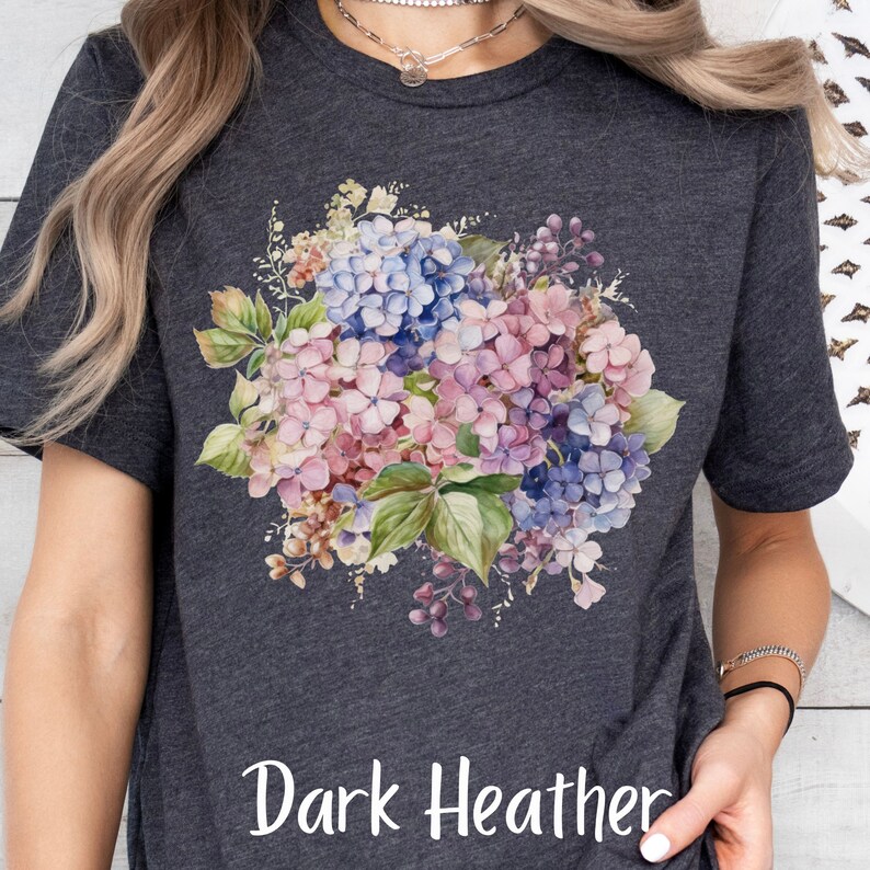 Peut inclure: Un t-shirt gris chin&eacute; fonc&eacute; avec un motif floral aquarelle repr&eacute;sentant des hortensias roses et bleus. Le texte "Dark Heather" est imprim&eacute; en bas du t-shirt.