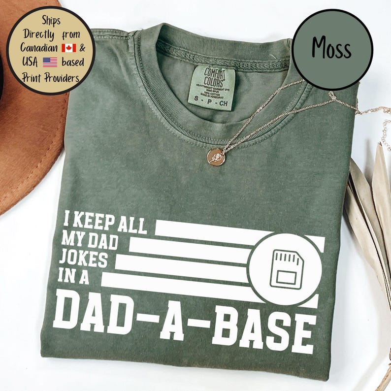 May include: Moss green t-shirt with the text "I KEEP ALL MY DAD JOKES IN A DAD-A-BASE" in white. The shirt features a graphic of a data storage icon. A gold necklace is visible.