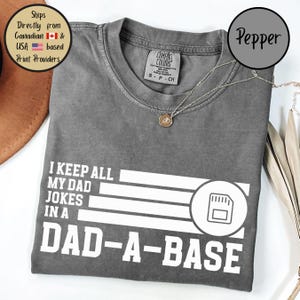 May include: Pepper-colored Comfort Colors t-shirt with the text "I KEEP ALL MY DAD JOKES IN A DAD-A-BASE" in white. The shirt has a round neck and short sleeves. A silver necklace with a pendant is on the shirt.