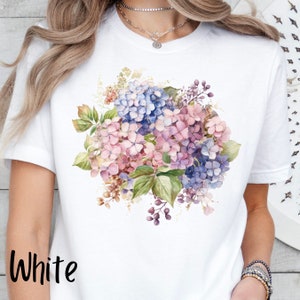 Peut inclure: T-shirt blanc avec un motif floral aquarelle de hortensias roses, bleus et violets.