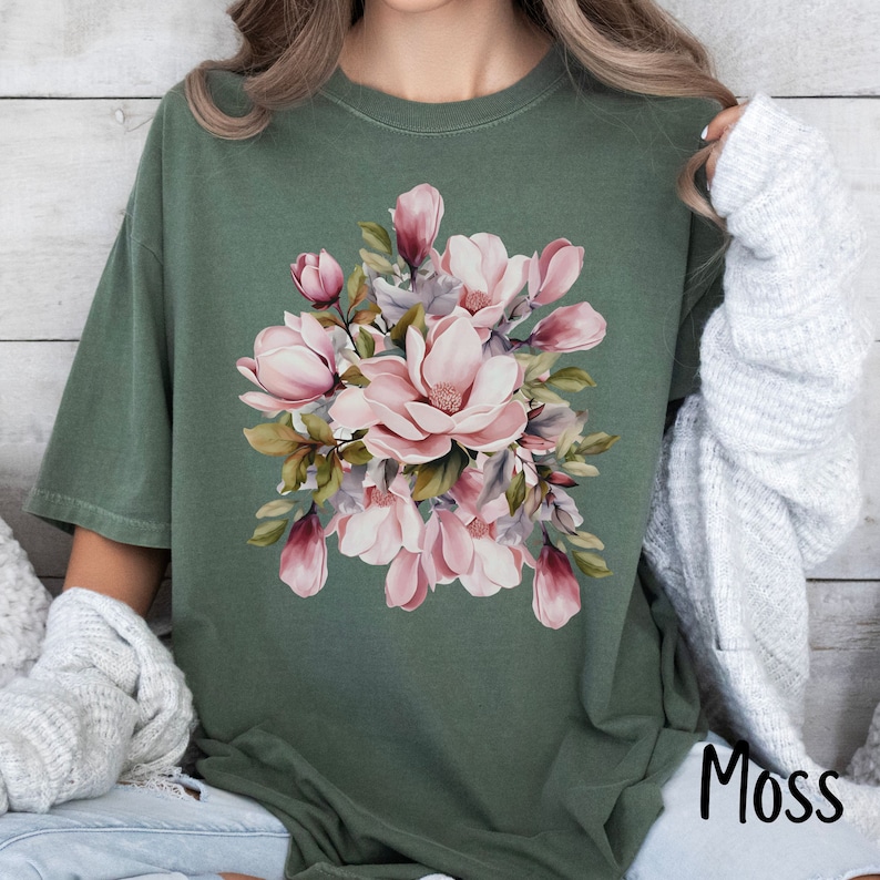 Peut inclure: Un t-shirt vert avec un motif floral compos&eacute; de fleurs de magnolia roses et blanches. Les fleurs sont dispos&eacute;es en bouquet avec des feuilles et des tiges vertes.