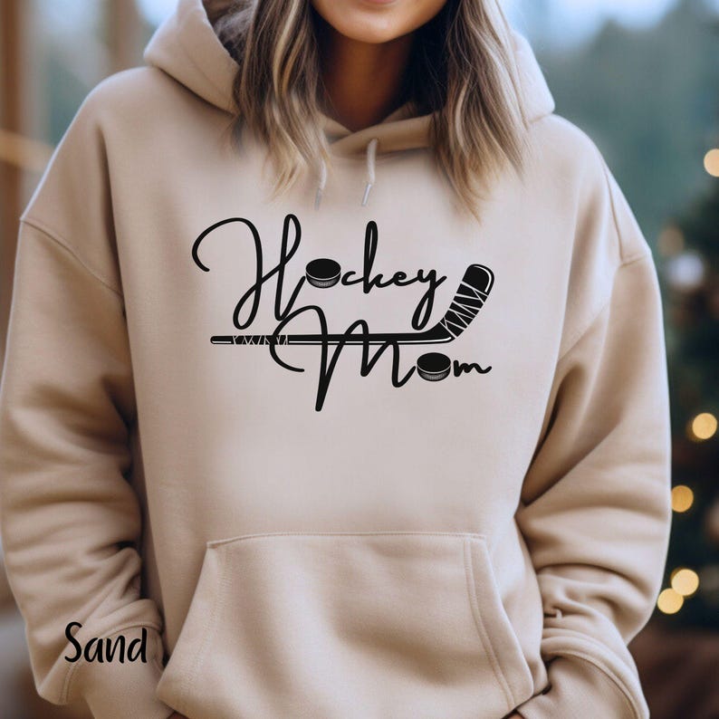 Op de afbeelding: Een zandkleurige hoodie met een zwarte afbeelding waarop "Hockey Mom" staat met een hockeystick en een puck.