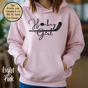 May include: Light pink hoodie with a front pocket and a black graphic that says "Hockey Girl" with a hockey puck and stick. The hoodie is made of a soft material and has a hood.