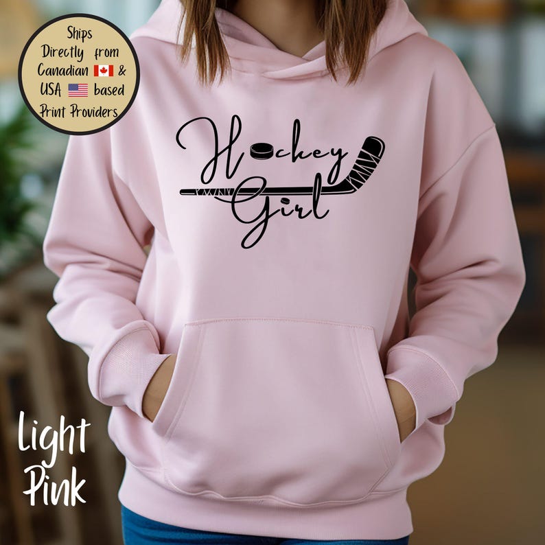 May include: Light pink hoodie with a front pocket and a black graphic that says "Hockey Girl" with a hockey puck and stick. The hoodie is made of a soft material and has a hood.