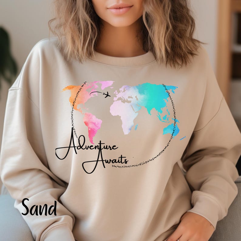 May include: A beige sweatshirt with a watercolor world map design and the text "Adventure Awaits". The map is in shades of pink, blue, and orange.