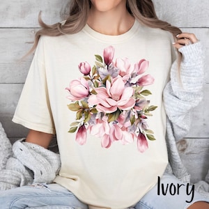 Peut inclure: Un t-shirt blanc avec un motif floral compos&eacute; de fleurs de magnolia roses et vertes. Le texte "Ivoire" est imprim&eacute; dans le coin inf&eacute;rieur droit du t-shirt.