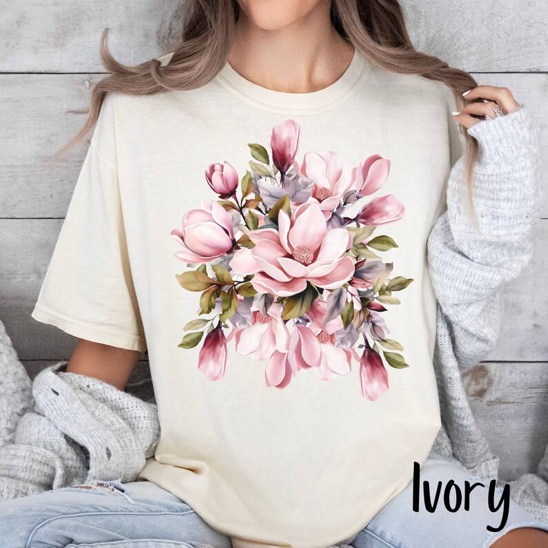 Peut inclure: Un t-shirt blanc avec un motif floral compos&eacute; de fleurs de magnolia roses et vertes. Le texte "Ivoire" est imprim&eacute; dans le coin inf&eacute;rieur droit du t-shirt.