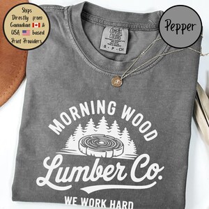 以下が含まれることがあります： 「Morning Wood Lumber Co. We Work Hard」と白で書かれたグレーのTシャツ。グラフィックには、成長輪のあるスタイリッシュな木の切り株と、背景に森が描かれています。