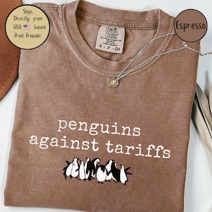 May include: A brown t-shirt with the text "penguins against tariffs" printed in white. A group of cartoon penguins are pictured below the text.