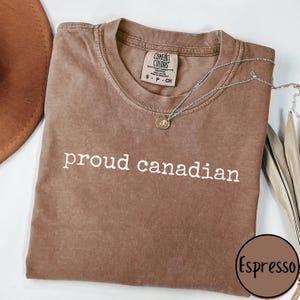 May include: A brown t-shirt with the text "proud canadian" printed in white. The shirt is made of a soft, comfortable material and has a distressed look. The shirt is shown on a flat surface with a brown hat and a silver necklace.