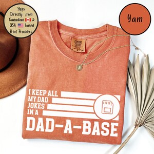 May include: A coral-colored t-shirt with the text "I KEEP ALL MY DAD JOKES IN A DAD-A-BASE" in white. The shirt has a crew neck and a small tag. A gold necklace with a pendant is draped across the shirt.