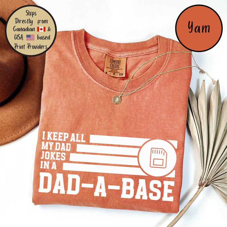 May include: A coral-colored t-shirt with the text "I KEEP ALL MY DAD JOKES IN A DAD-A-BASE" in white. The shirt has a crew neck and a small tag. A gold necklace with a pendant is draped across the shirt.