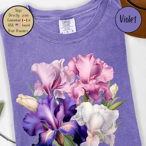 May include: A violet-colored t-shirt featuring a floral design of irises in shades of purple, pink, and white. The shirt has a crew neck and a Comfort Colors label. The word "Violet" is in a circle on the upper right.