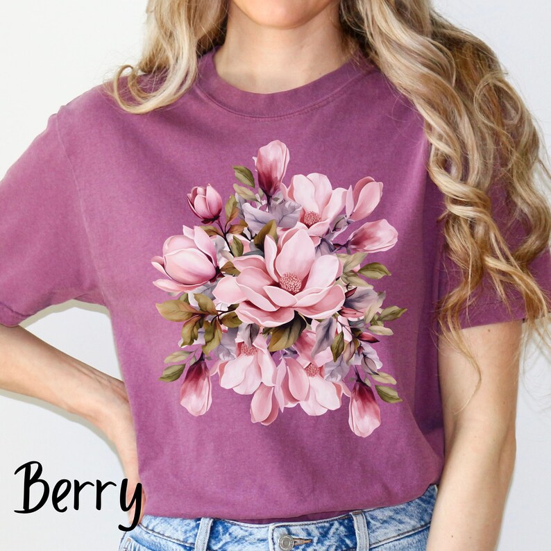 Peut inclure: Un t-shirt mauve avec un imprim&eacute; floral de fleurs de magnolia roses et blanches avec des feuilles vertes. Le mot "Berry" est imprim&eacute; en noir en bas &agrave; gauche de l'image.