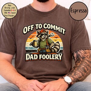 May include: Dark brown t-shirt featuring the text "OFF TO COMMIT DAD FOOLERY" and a raccoon graphic. The raccoon is wearing a red cap, sunglasses, and holding a wrench and beer. The shirt has a vintage, faded appearance.