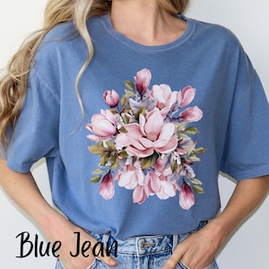 Peut inclure: Un t-shirt bleu jean avec un imprim&eacute; floral de fleurs de magnolia roses et blanches. Les fleurs sont dispos&eacute;es en bouquet avec des feuilles vertes.