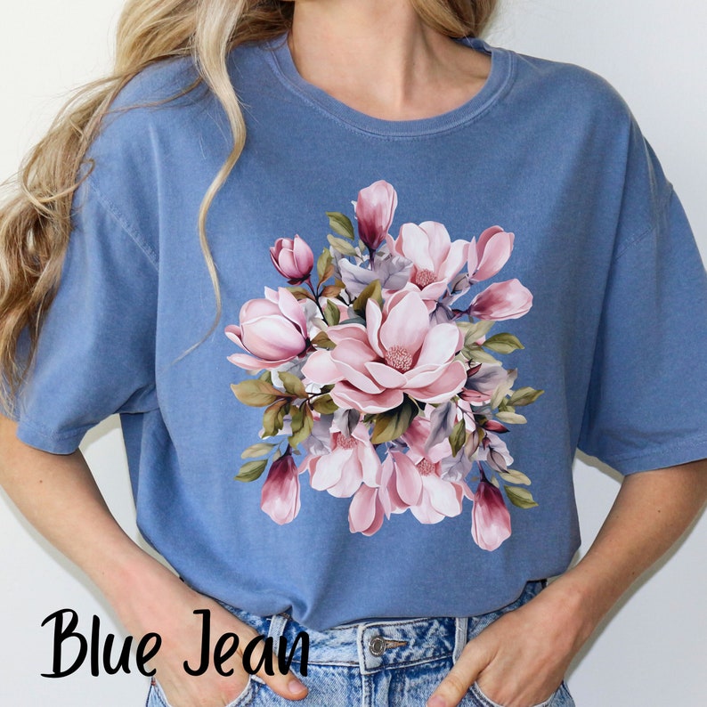 Peut inclure: Un t-shirt bleu jean avec un imprim&eacute; floral de fleurs de magnolia roses et blanches. Les fleurs sont dispos&eacute;es en bouquet avec des feuilles vertes.