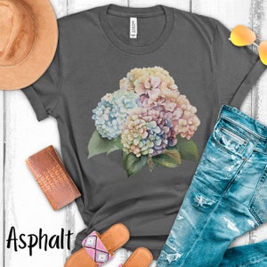 May include: A gray t-shirt with a watercolor floral design featuring a bouquet of hydrangeas in shades of blue, pink, and white. The shirt is laid flat on a white surface with a pair of sandals, a hat, and a wallet.