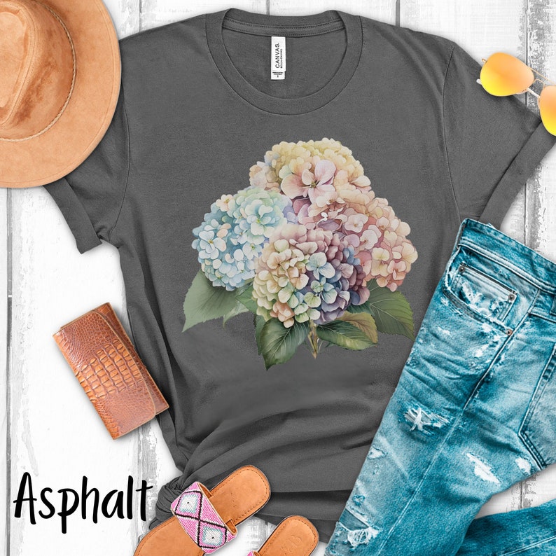 May include: A gray t-shirt with a watercolor floral design featuring a bouquet of hydrangeas in shades of blue, pink, and white. The shirt is laid flat on a white surface with a pair of sandals, a hat, and a wallet.