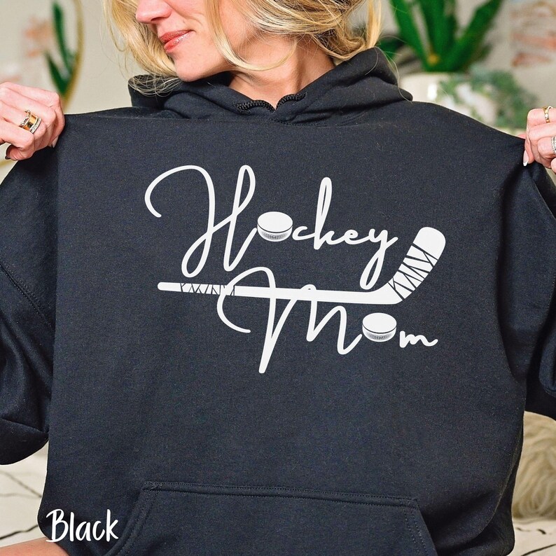 Op de afbeelding: Zwarte hoodie met witte tekst "Hockey Mom" met een afbeelding van een hockeystick en een puck.