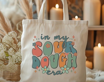 May include: A white canvas tote bag with the text "in my sourdough era" in a retro, colorful font. The text is surrounded by small flowers and stars.
