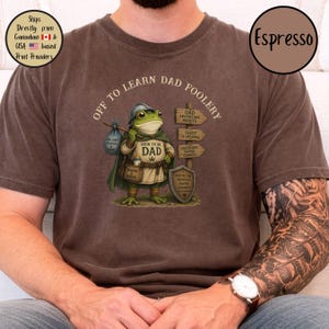 May include: Espresso-colored t-shirt with the phrase "Off to Learn Dad Foolery" and a cartoon frog dressed as a knight. The frog carries a bag labeled "Dad Jokes & Diapers & Love" and a sign that says "Soon to be Dad".