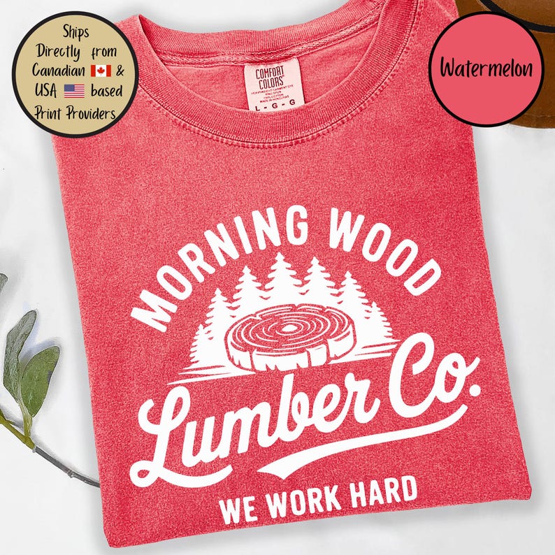 以下が含まれることがあります： 「Morning Wood Lumber Co. We Work Hard」と白で書かれた赤いTシャツ。グラフィックには、成長輪のあるスタイリッシュな木の切り株と、その背後に森が描かれている。