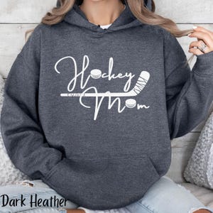 Op de afbeelding: Donkergrijze hoodie met een witte afbeelding met de tekst "Hockey Mom" met een hockeystick en een puck.