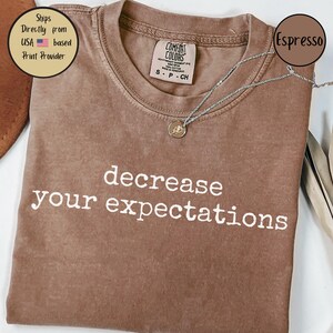 May include: A brown t-shirt with the text "decrease your expectations" printed in white. The shirt is made of a soft, comfortable material.