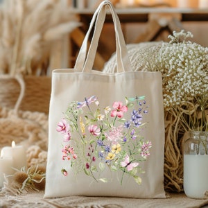 May include: A white canvas tote bag with a floral design featuring pink, yellow, and purple flowers, green leaves, and dragonflies. The bag has a natural fabric texture and two handles.