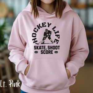 Op de afbeelding: Een roze hoodie met een zwarte afbeelding van een hockeyer in een cirkel met de tekst "Hockey Life" en "Skate, Shoot Score" onder de cirkel.