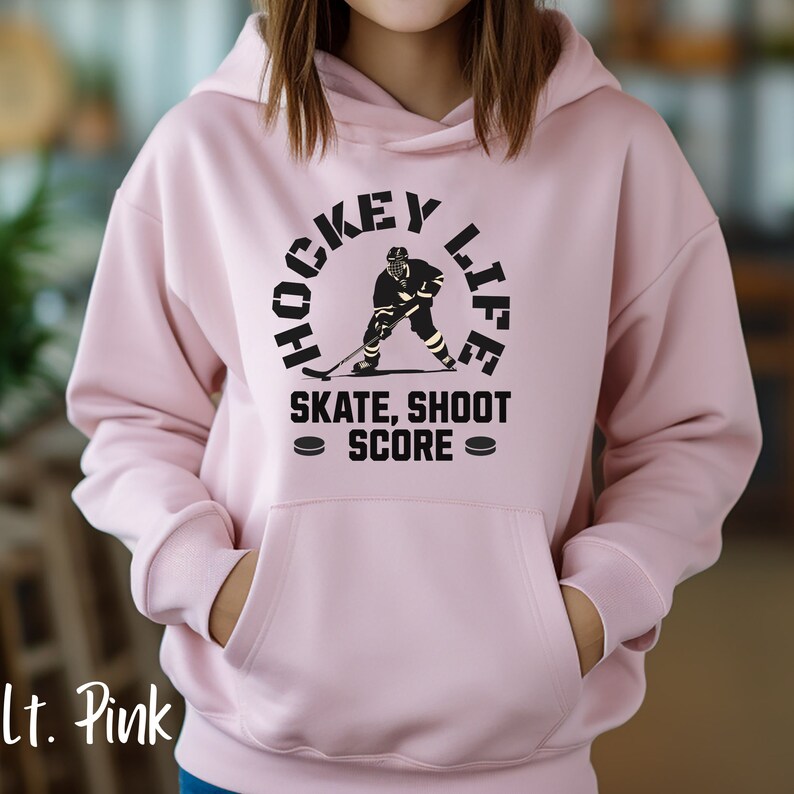 Op de afbeelding: Een roze hoodie met een zwarte afbeelding van een hockeyer in een cirkel met de tekst "Hockey Life" en "Skate, Shoot Score" onder de cirkel.