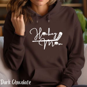 Op de afbeelding: Een donkerbruine hoodie met een witte afbeelding waarop "Hockey Mom" staat met een hockeystick en een puck.
