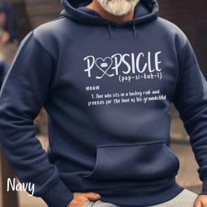 May include: A navy blue hoodie with a white graphic that says "Popsicle (pop-si-kuh-1)" and defines the word as "One who sits in a hockey rink and freezes for the love of his grandchild".