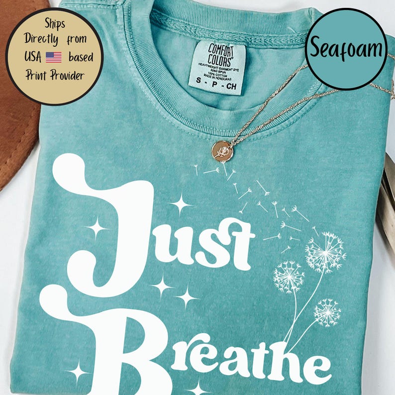 May include: Seafoam green t-shirt featuring the words "Just Breathe" in white script and a dandelion graphic. The shirt has a crew neck and short sleeves. A gold necklace with a small pendant is visible.