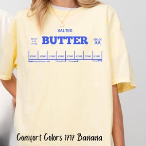 May include: A yellow t-shirt with the words "SALTED BUTTER GRADE AA" printed in blue. The shirt also has a blue ruler with measurements in tablespoons and cups.