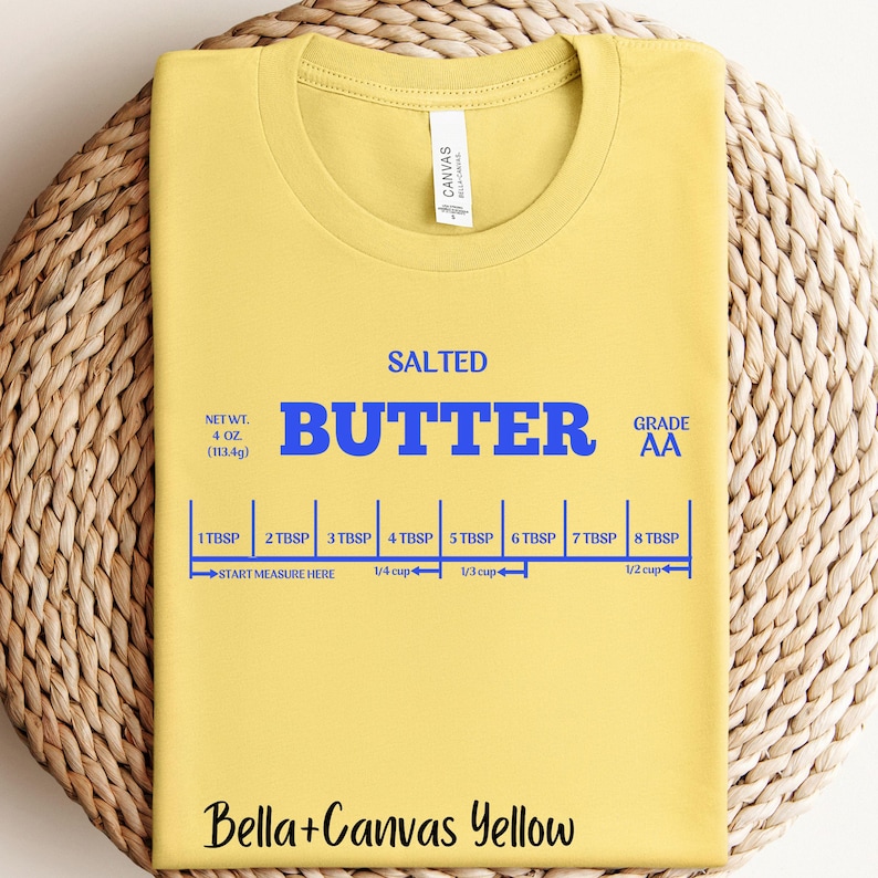 May include: A yellow Bella+Canvas t-shirt with a graphic of a stick of butter with the text "SALTED BUTTER GRADE AA" and a measurement guide in tablespoons and cups. The text "NET WT. 4 OZ (113.4g)" is also included.