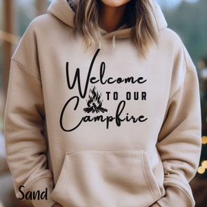 Peut inclure: Un sweat à capuche beige avec un motif noir qui dit "Bienvenue à notre feu de camp" avec une illustration de feu de camp.