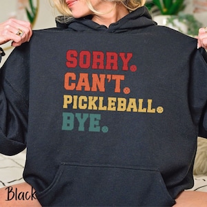 Peut inclure: Sweat-shirt à capuche noir avec le texte "SORRY. CAN'T. PICKLEBALL. BYE." en lettres rétro colorées avec des icônes de pickleball.