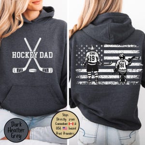 May include: Dark heather grey hoodie with the text "HOCKEY DAD" and crossed hockey sticks and a puck. The back features an American flag design with hockey players wearing the numbers 19 and 91. The hoodie has a hood and a front pocket.