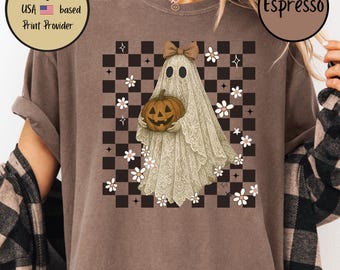 May include: Espresso-colored t-shirt featuring a Halloween-themed graphic. The design includes a ghost wearing a bow and holding a carved pumpkin, set against a checkered background with floral accents. The shirt is a comfortable, casual wear.