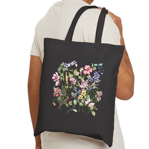 May include: A black tote bag with a floral design featuring pink, purple, yellow, and blue flowers, green leaves, and dragonflies. The bag has a black canvas material and two black straps.