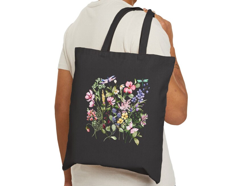 May include: A black tote bag with a floral design featuring pink, purple, yellow, and blue flowers, green leaves, and dragonflies. The bag has a black canvas material and two black straps.