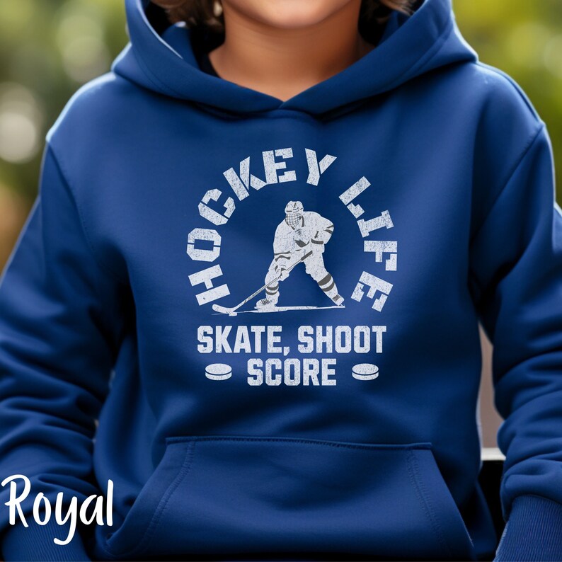 Op de afbeelding: Koninklijk blauwe hoodie met een witte afbeelding van een hockeyer in een cirkel met de tekst "Hockey Life" en "Skate, Shoot Score" eronder.