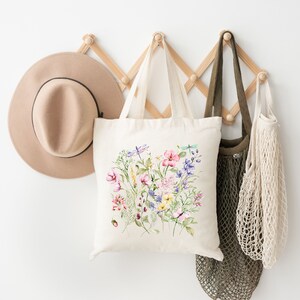 May include: A white canvas tote bag with a floral design featuring pink, purple, yellow, and blue flowers, green leaves, and dragonflies. The bag is hanging on a wooden rack with a brown hat and a mesh bag.