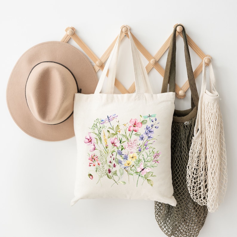 May include: A white canvas tote bag with a floral design featuring pink, purple, yellow, and blue flowers, green leaves, and dragonflies. The bag is hanging on a wooden rack with a brown hat and a mesh bag.
